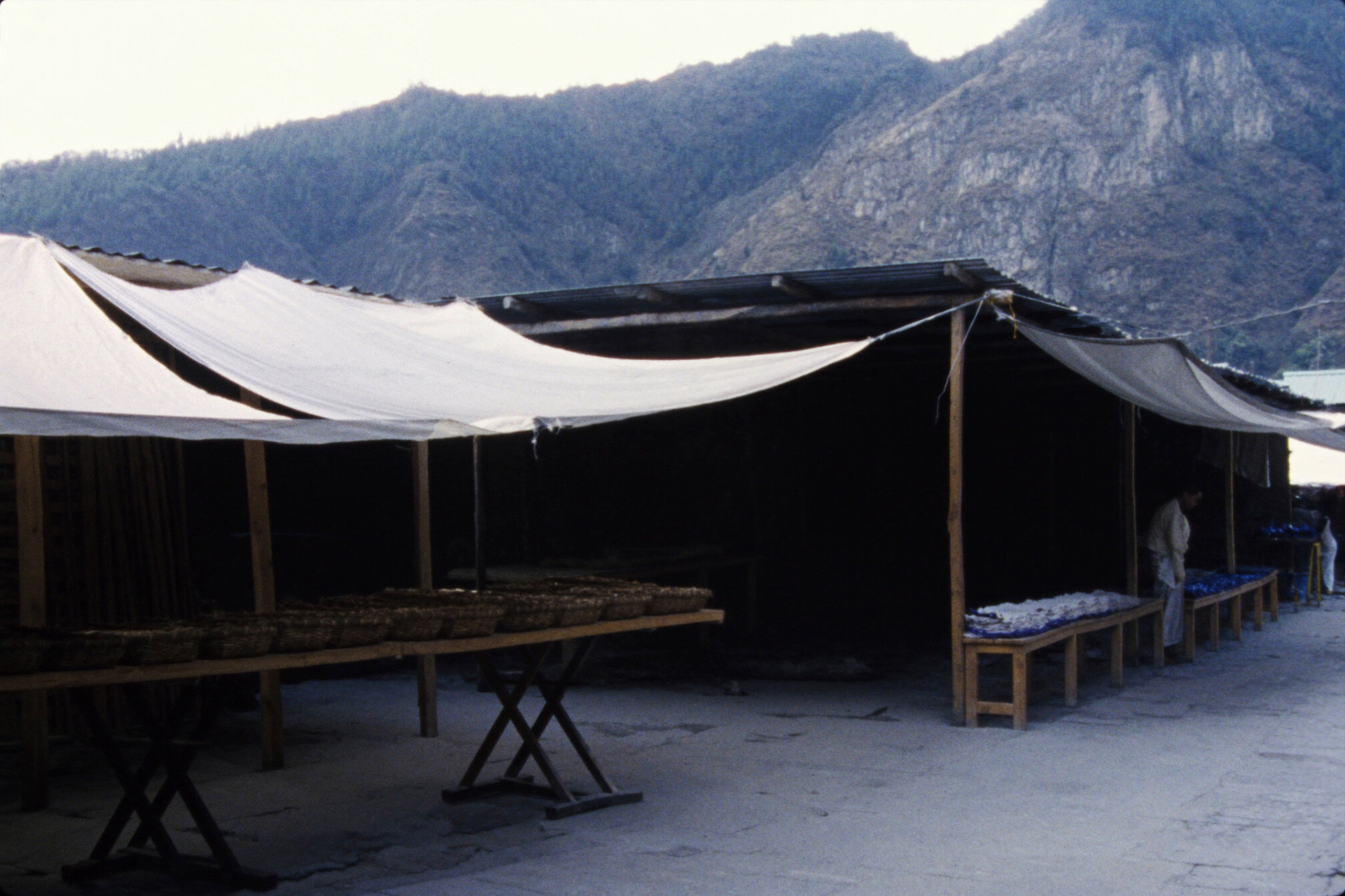 An empty open-air market street in a Himalayan town, timber stalls under canvas awnings and a mountain ridge above the rooftops