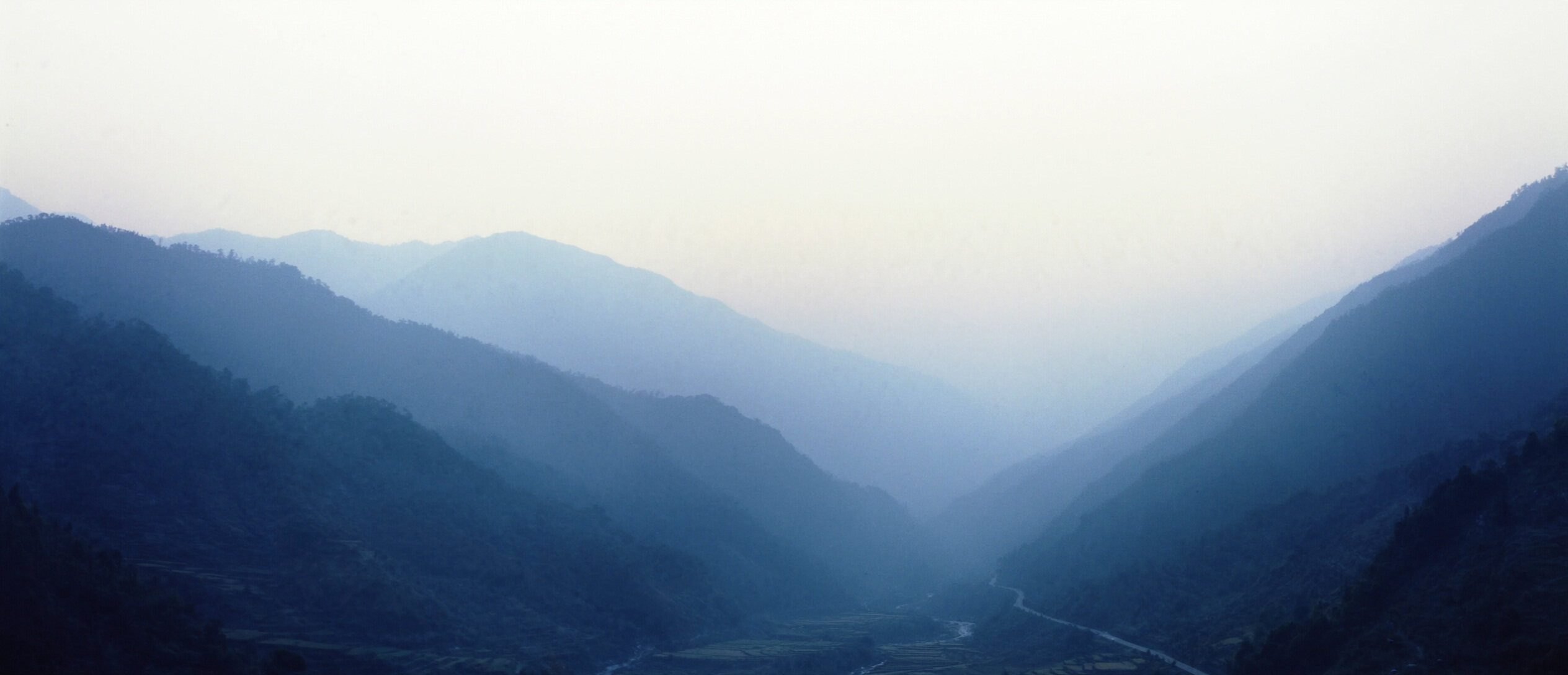 A highland valley under soft morning haze, terraced fields on the valley floor and a single road running out toward the horizon
