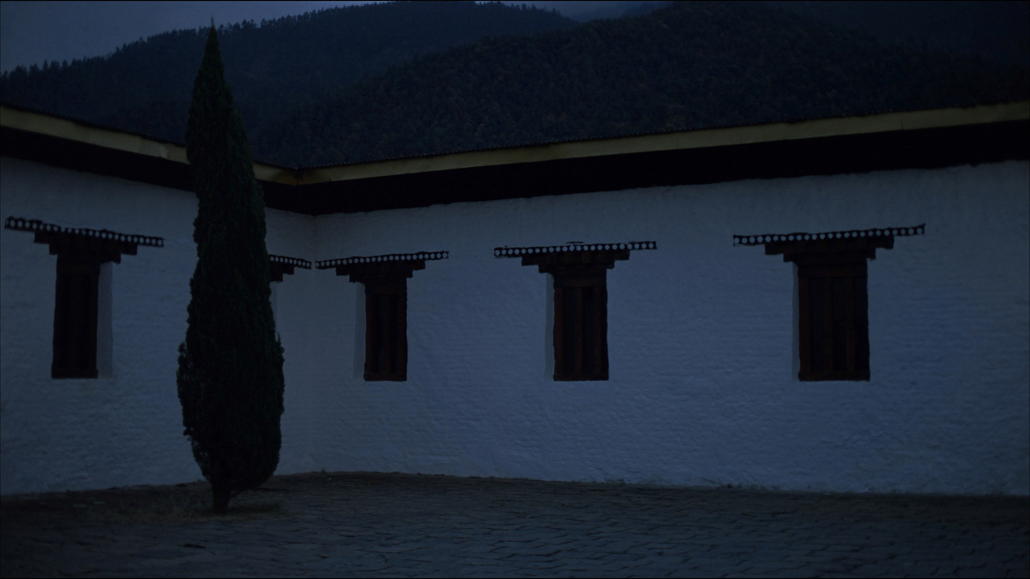 Whitewashed Bhutanese walls with dark timber windows and a gilded roof edge, a forested ridge fading into mist behind