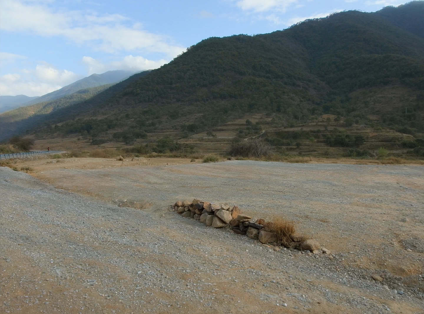 An empty building plot beside a hillside highway, cleared ground with a stone boundary and a forested ridge beyond