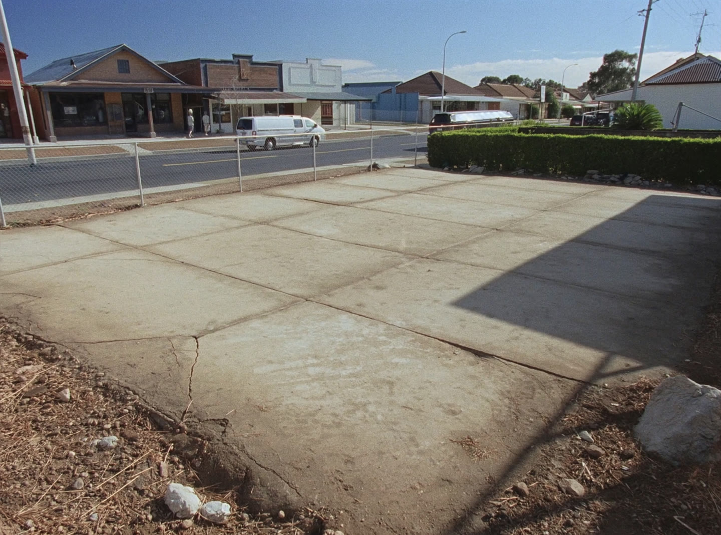 An empty commercial corner lot in an Australian suburb with a concrete hardstand and shops across the road