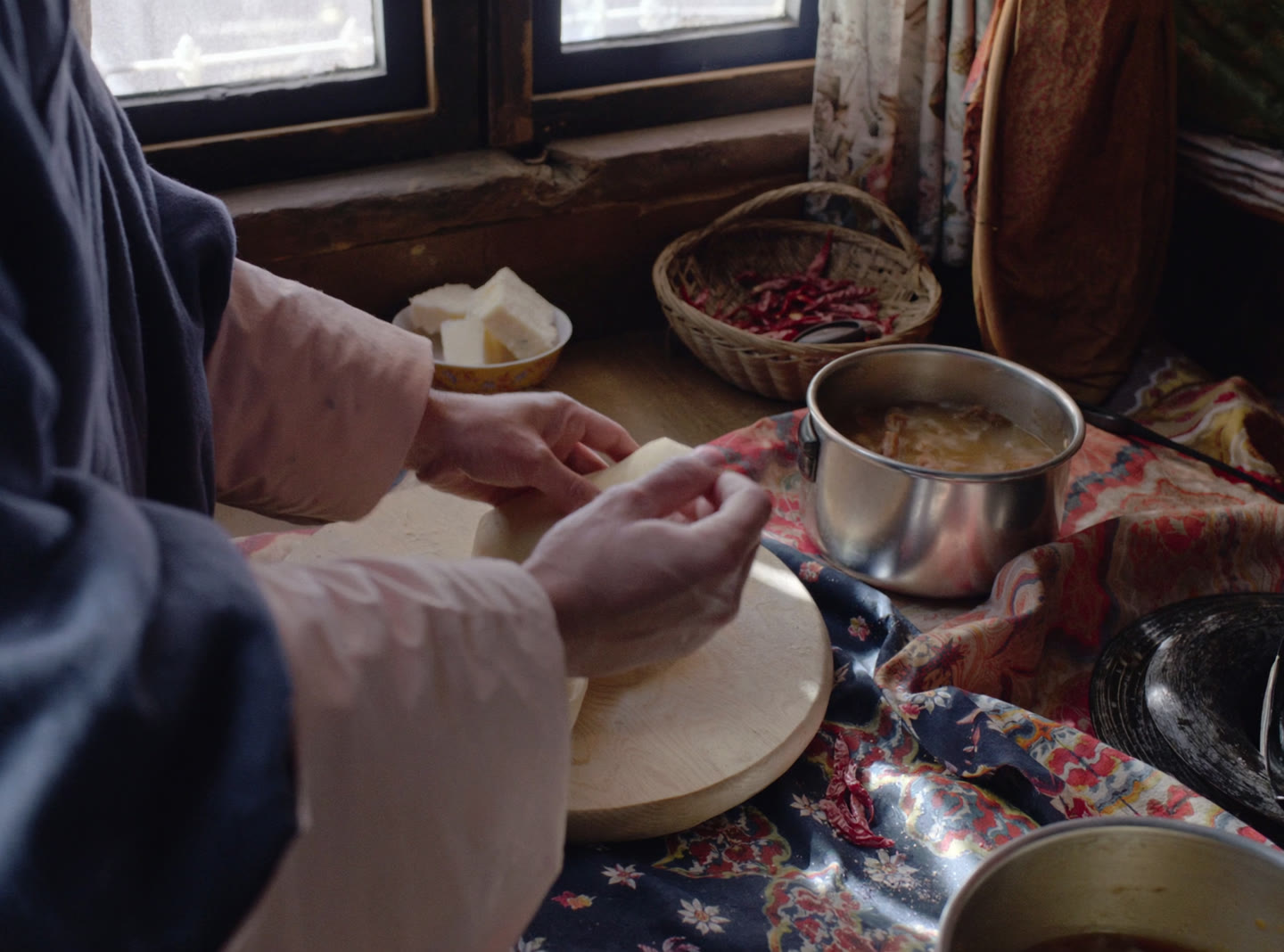 Hands shaping dough on a wooden board beside a pot of stew and a basket of dried chillies
