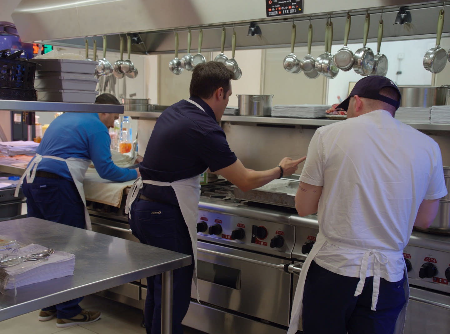 Students in white aprons working at a stainless range while an instructor points into a pan in a training kitchen