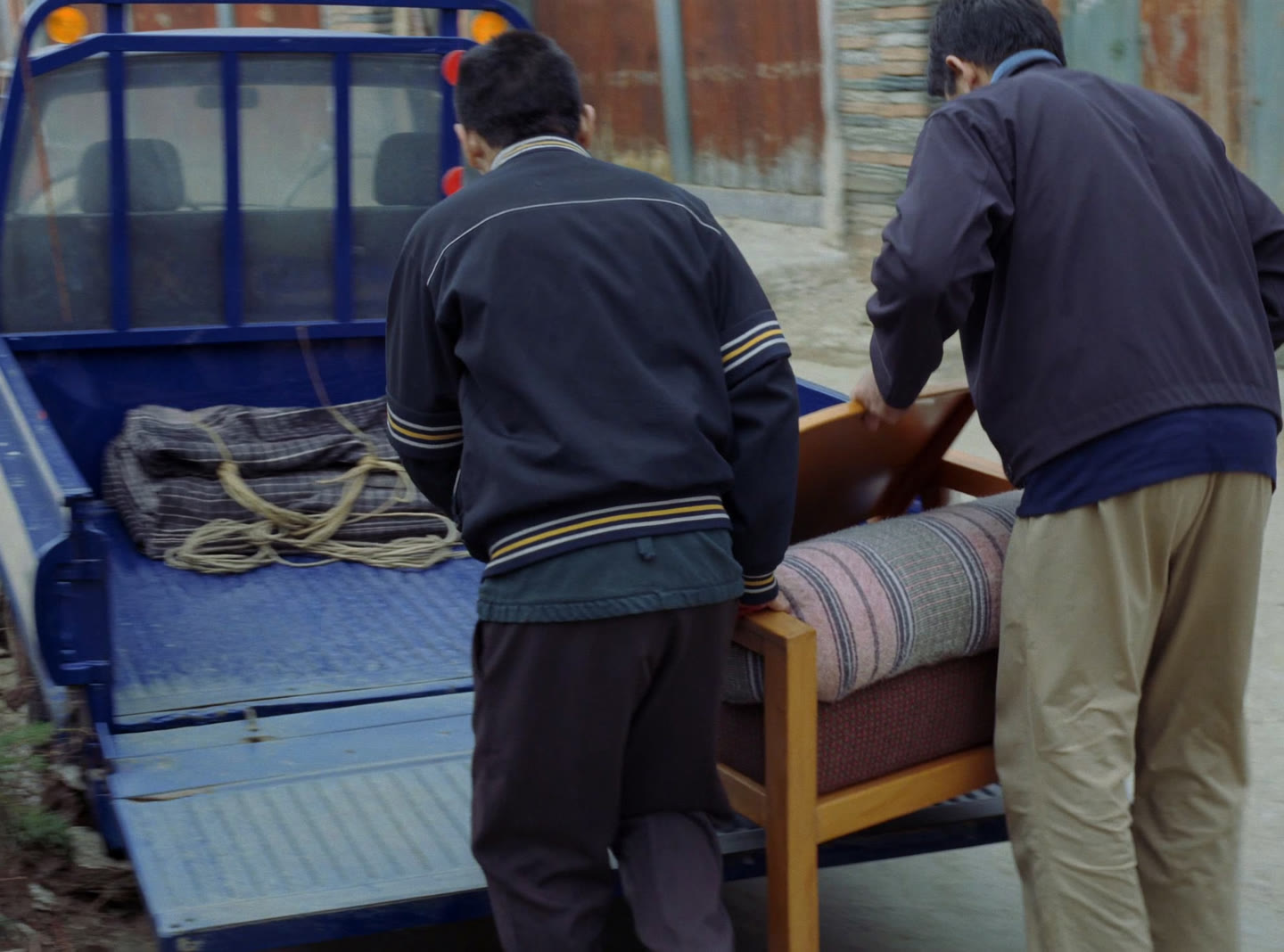 Two movers carrying an armchair down the ramp of a small blue truck on a quiet street