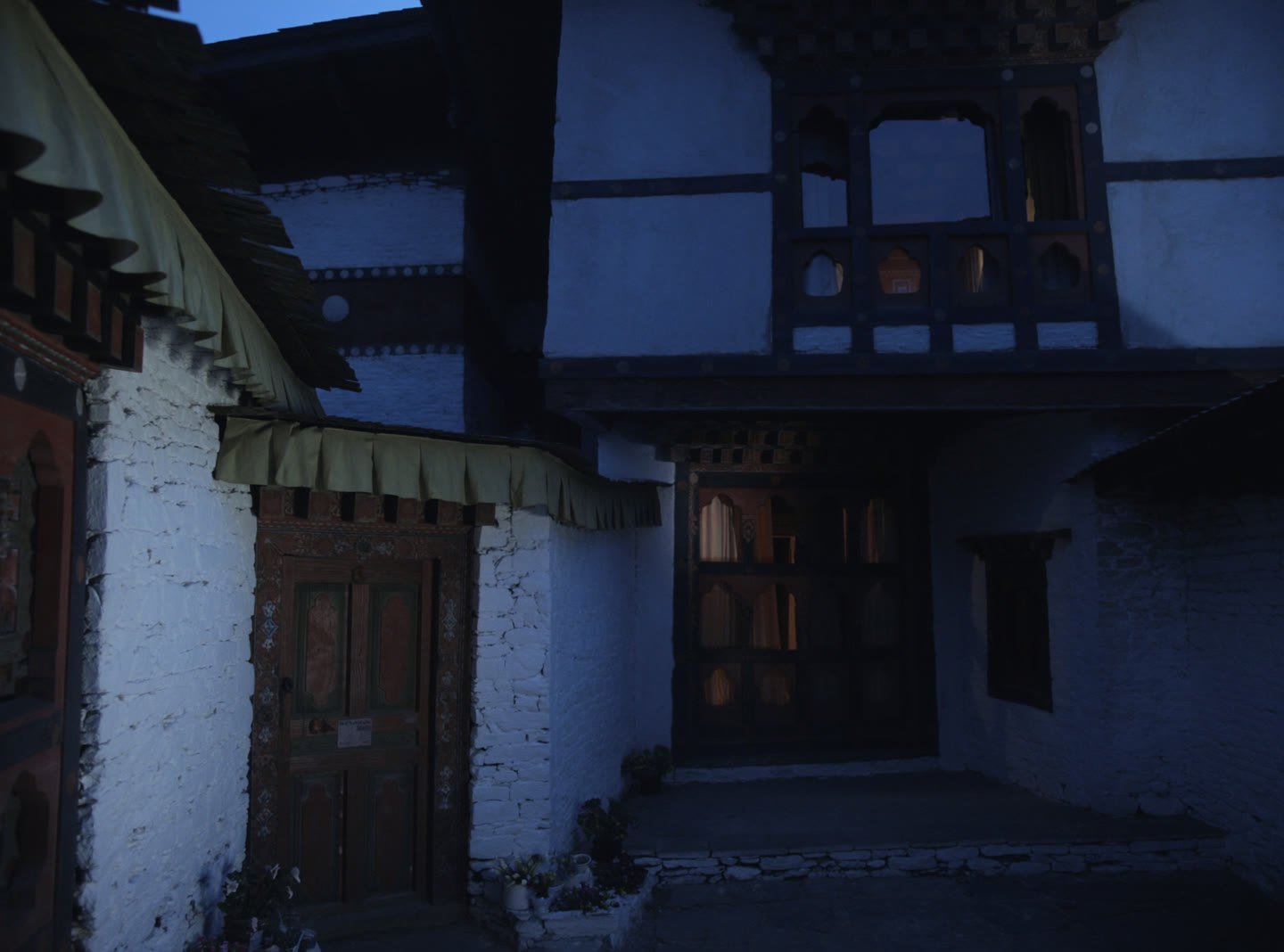 A modest Bhutanese guesthouse at dusk, whitewashed walls, carved timber entrance and warm lit windows