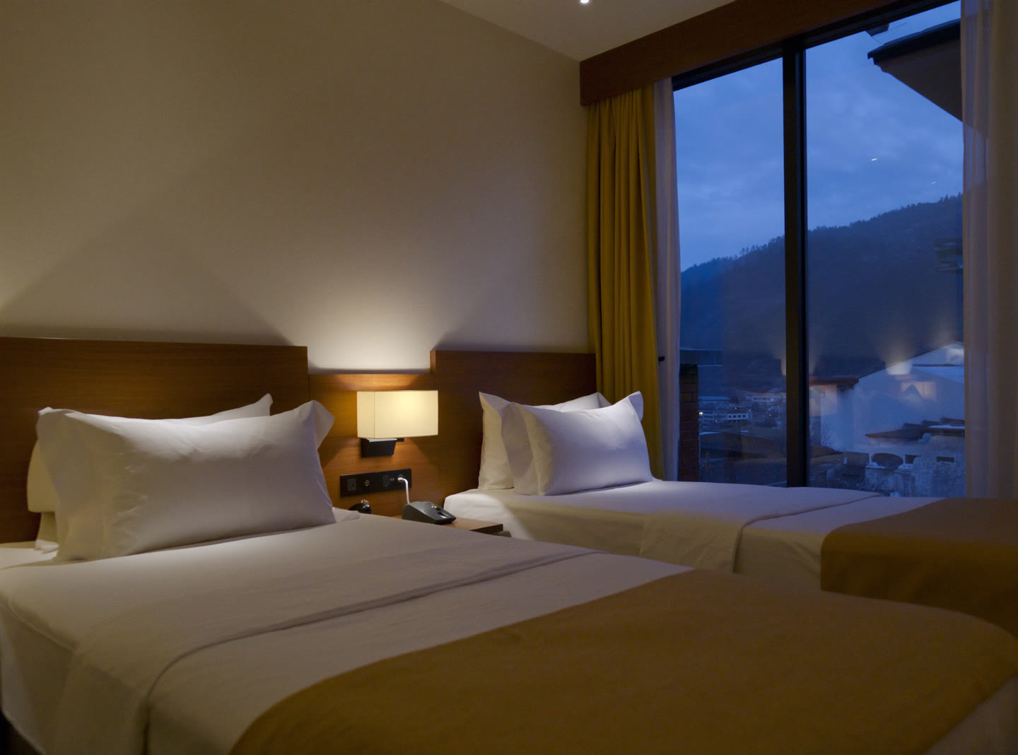 A modern twin hotel room in Thimphu, two made single beds and a lit bedside lamp at dusk