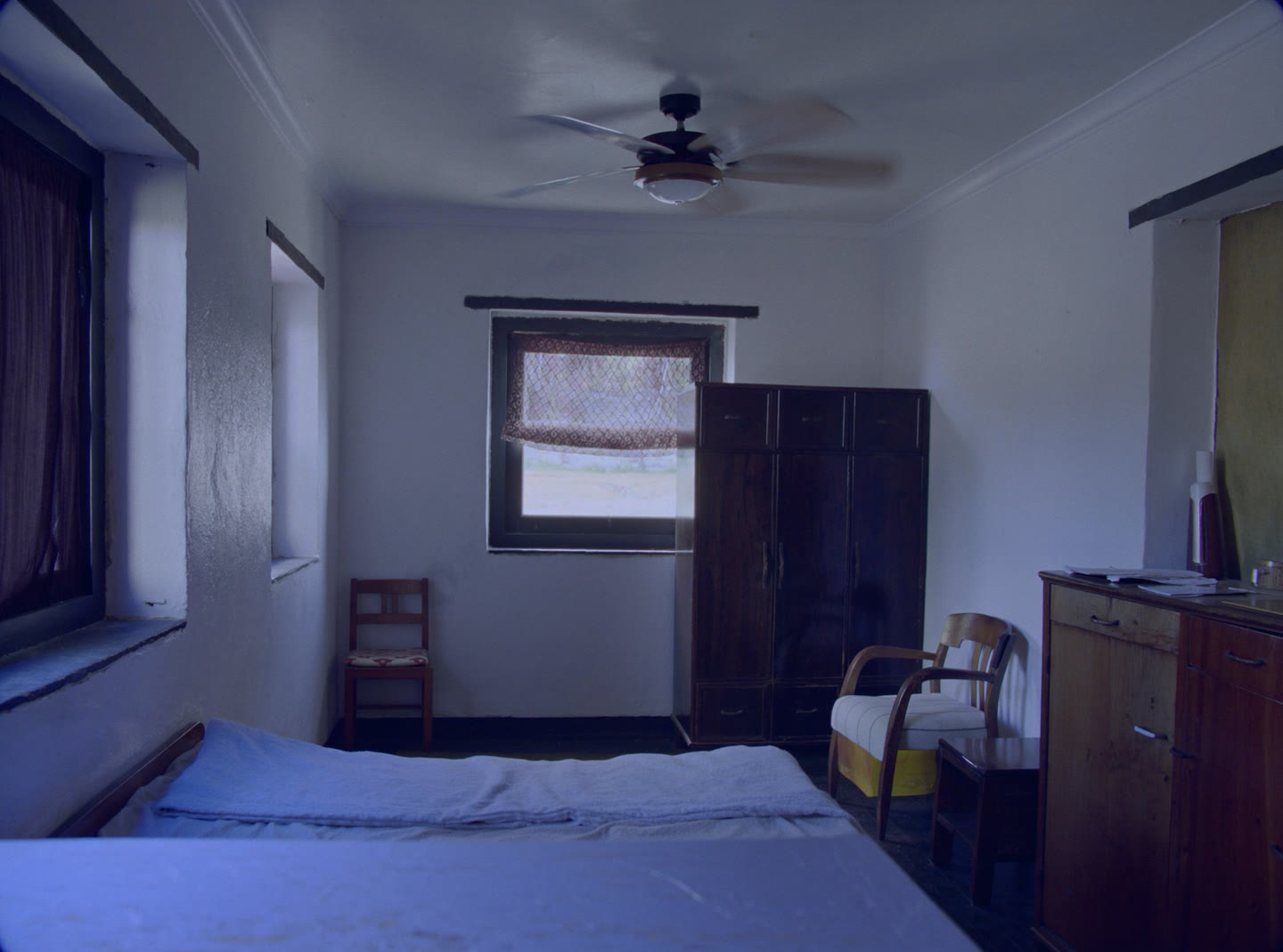 A simple furnished single room to let, a bed, a small desk and a net-curtained window in Bhutanese daylight
