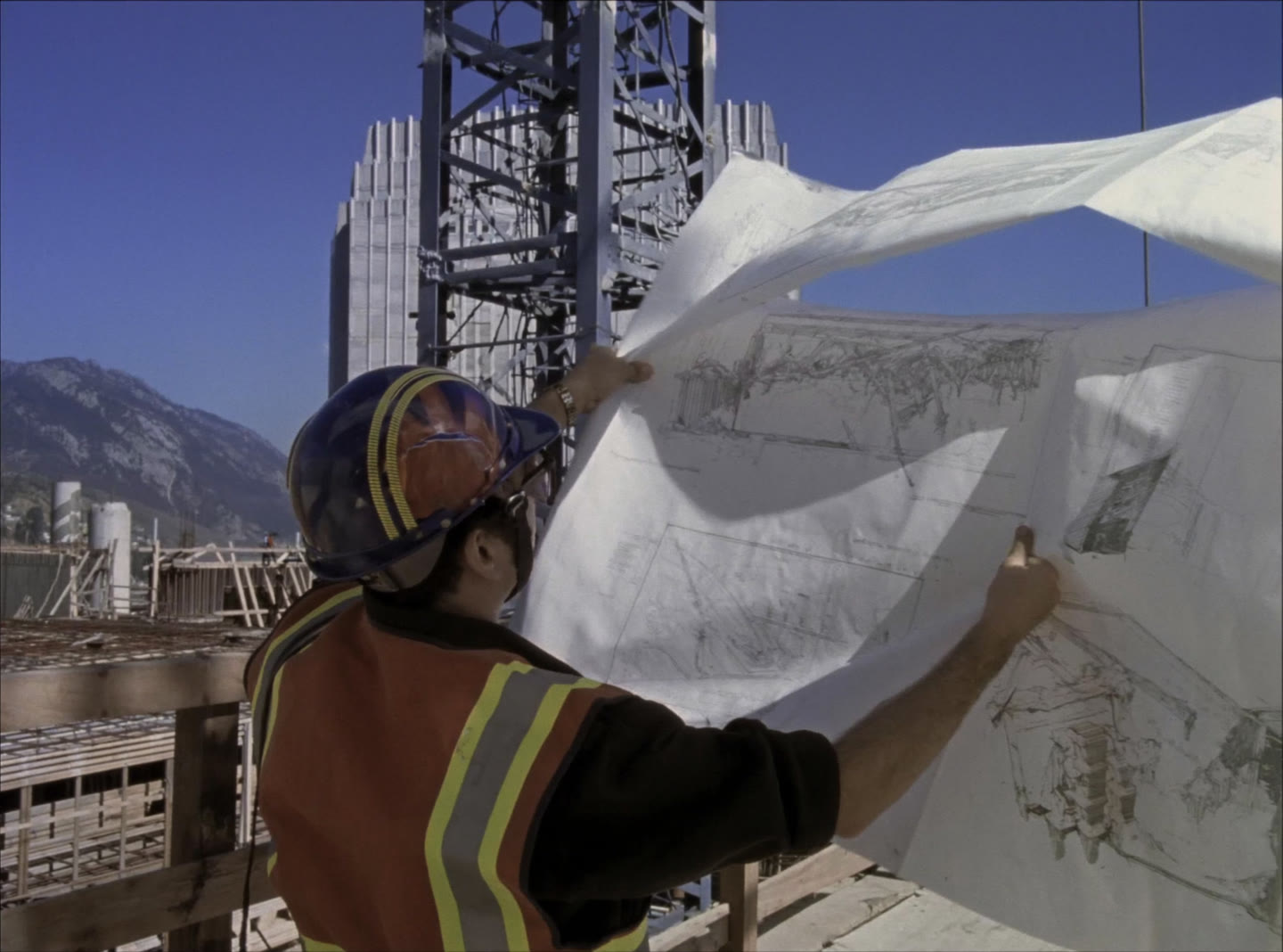 A site supervisor in a hard hat and high-visibility vest on a high-rise deck holding unrolled drawings