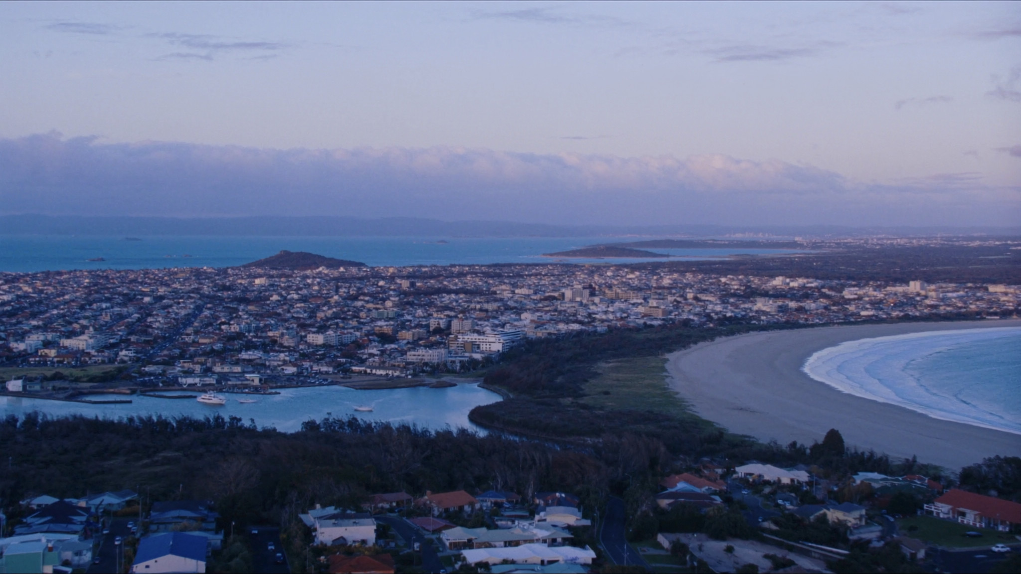 An elevated view over an Australian coastal city in the late afternoon, a pale beach curving along the shore